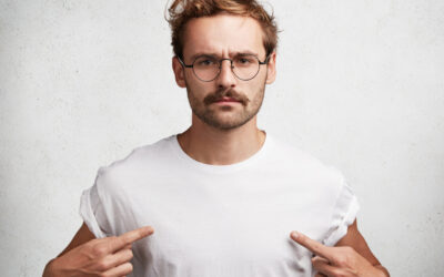 Photo of serious confident fashionable male with trendy hairdo, wears round spectacles and white t shirt, indicates at blank copy space of his clothes for your designing content. People and clothing