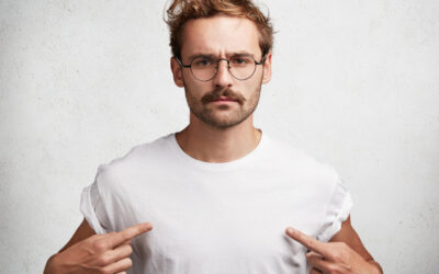 Photo of serious confident fashionable male with trendy hairdo, wears round spectacles and white t shirt, indicates at blank copy space of his clothes for your designing content. People and clothing