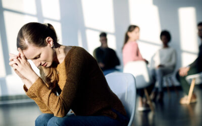 Young depressed woman struggling with herself while sitting apart from participants of group therapy.
